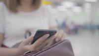 Glückliche Asiatin, die ihren Smartphone beim Sitzen auf Stuhl in der Abfertigungshalle beim Warten ihres Fluges am Abfluggatter im internationalen Flughafen verwendet und überprüft.