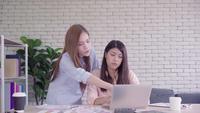 Zwei junge asiatische Frauen, die zusammen in einem Büro an ihrem Kleinbetrieb sitzen, einen Bericht oder eine Schreibarbeit mit erfreutem Lächeln lesend.